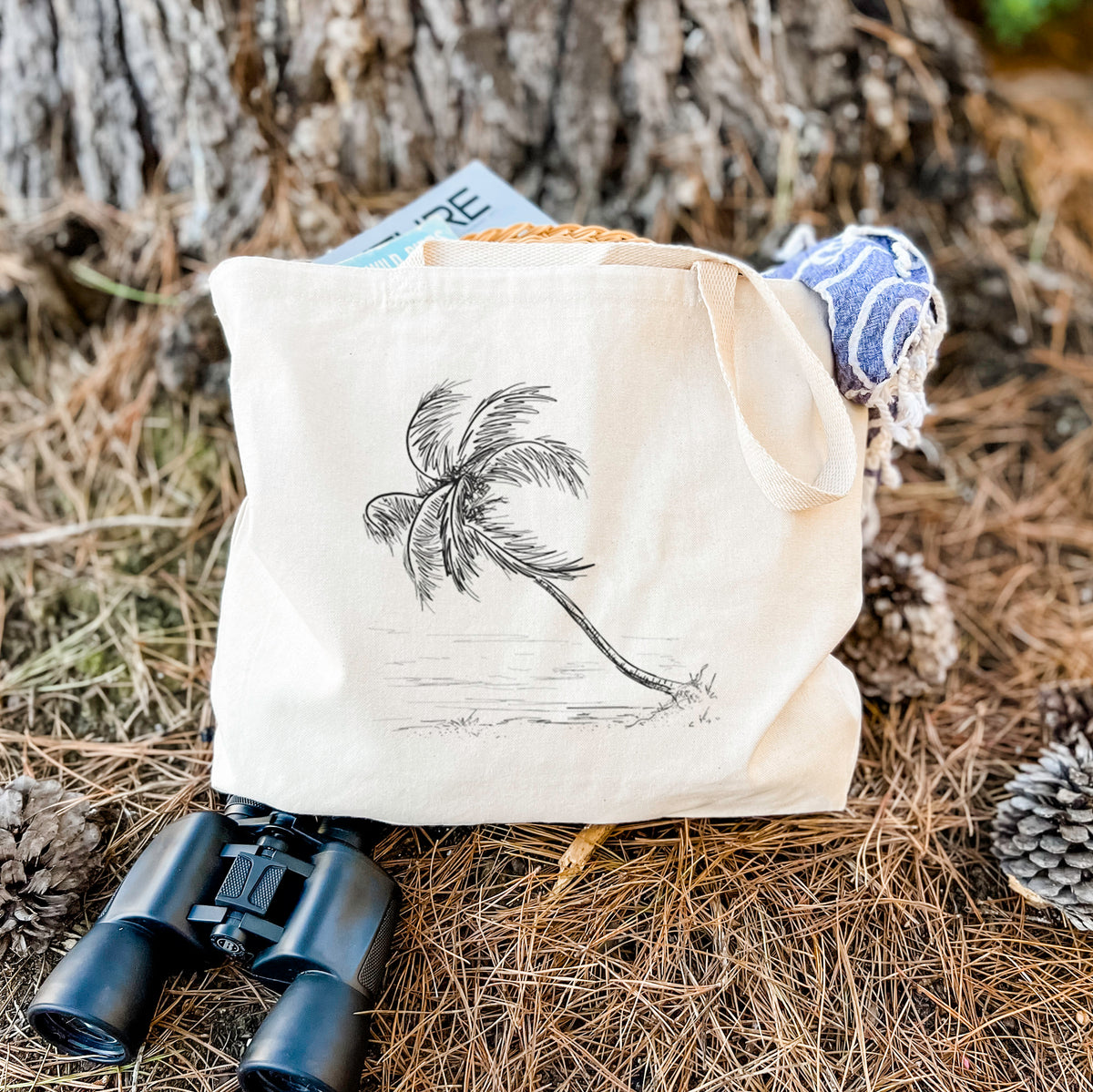 Canvas tote bag printed with a black ink illustration of a coconut palm leaning over a sandy beach, resting on dry grass beside a pair of binoculars.