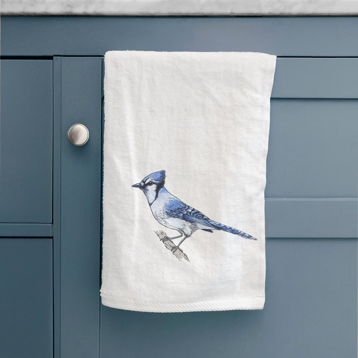 White hand towel printed with a full-color illustration of a blue jay perched on a branch, hanging on a cabinet door.