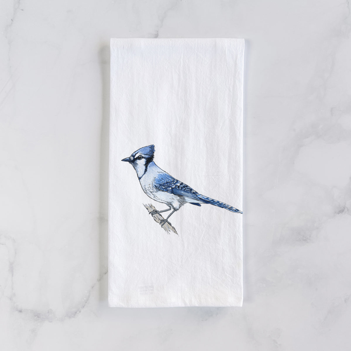 White cotton tea towel laid flat on marble, printed with a full-color illustration of a blue jay perched on a branch.