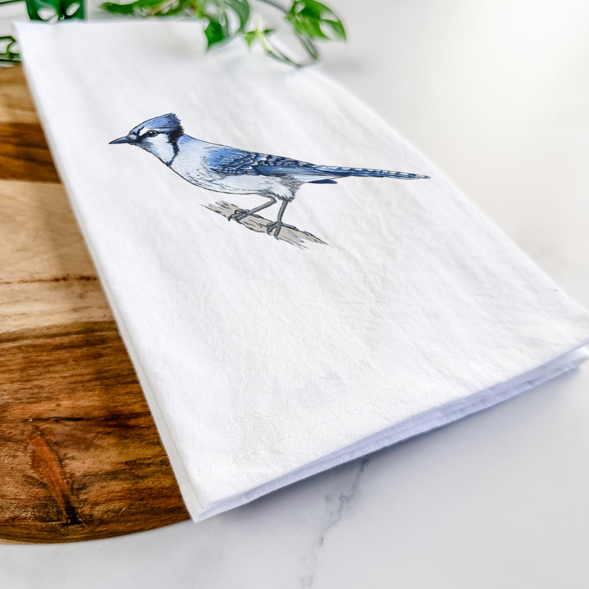 Close-up of a folded white tea towel on a wooden board, showing a full-color illustration of a blue jay perched on a branch.