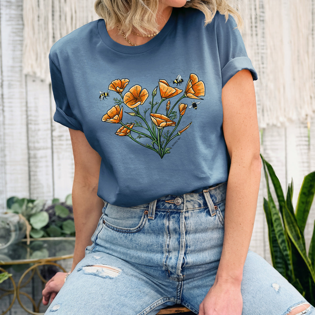 Woman wearing a steel blue lightweight cotton t-shirt printed with a full-color bouquet of orange California poppies arranged in a heart shape, with small bees.