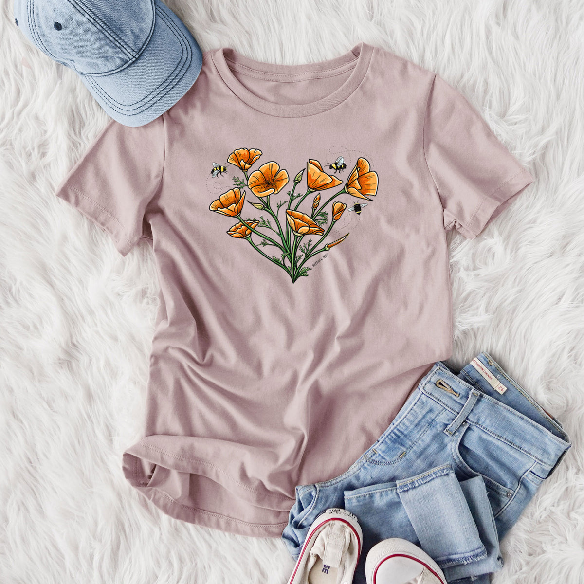 Women's relaxed fit cotton t-shirt printed with a full-color bouquet of orange California poppies arranged in a heart shape, with small bees, styled flat on a white faux fur rug with jeans.