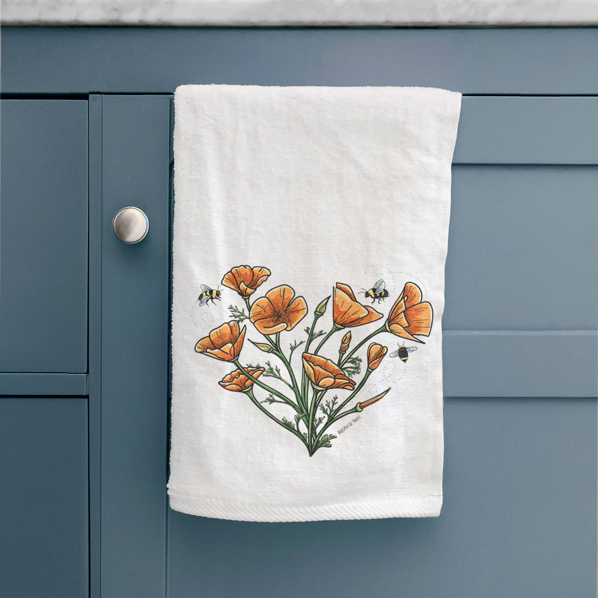 White hand towel printed with a full-color bouquet of orange California poppies arranged in a heart shape, with small bees, hanging on a cabinet door.