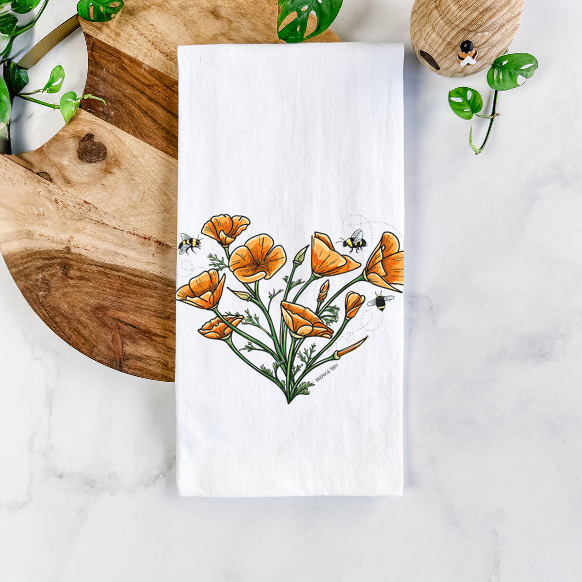 White cotton tea towel printed with a full-color bouquet of orange California poppies arranged in a heart shape, with small bees, on a marble counter beside a round wooden cutting board and greenery.