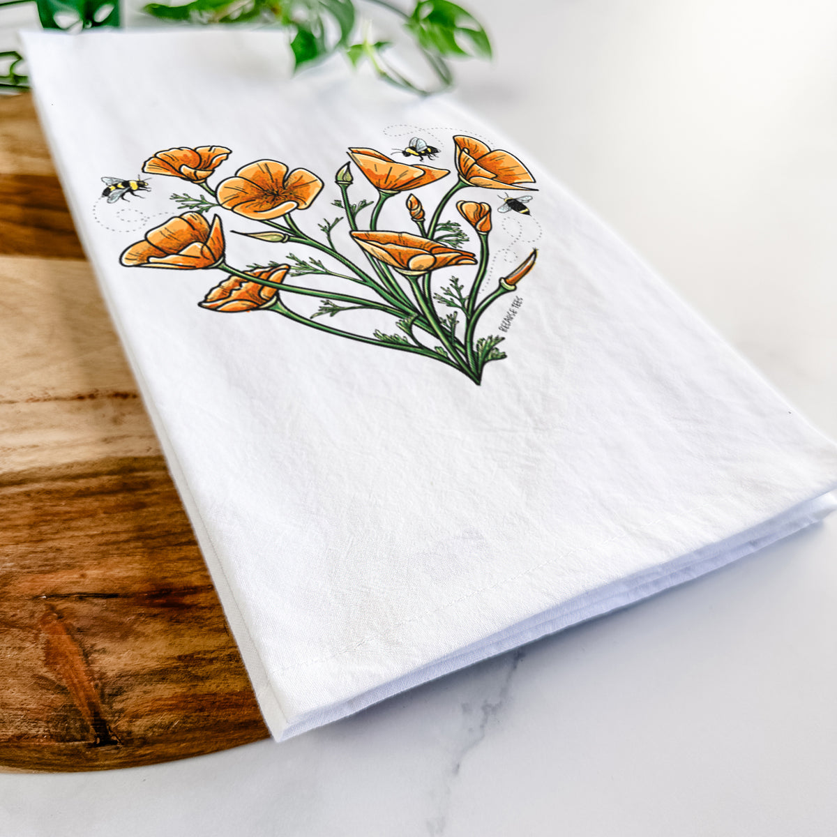 Close-up of a folded white tea towel on a wooden board, showing a full-color bouquet of orange California poppies arranged in a heart shape, with small bees.