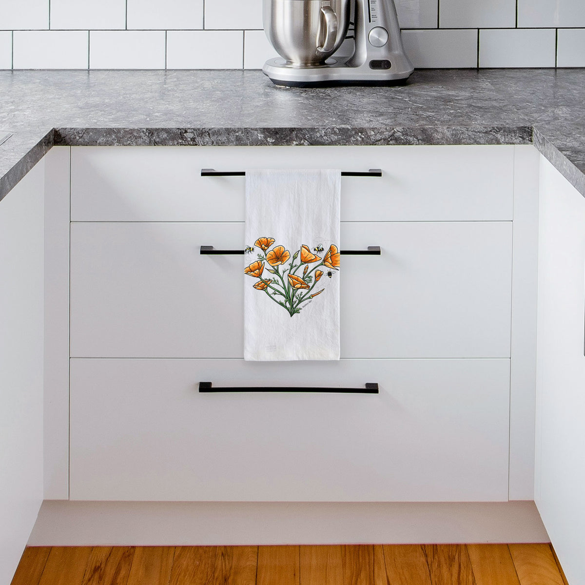 White tea towel printed with a full-color bouquet of orange California poppies arranged in a heart shape, with small bees, hanging from a drawer handle in a bright white kitchen.