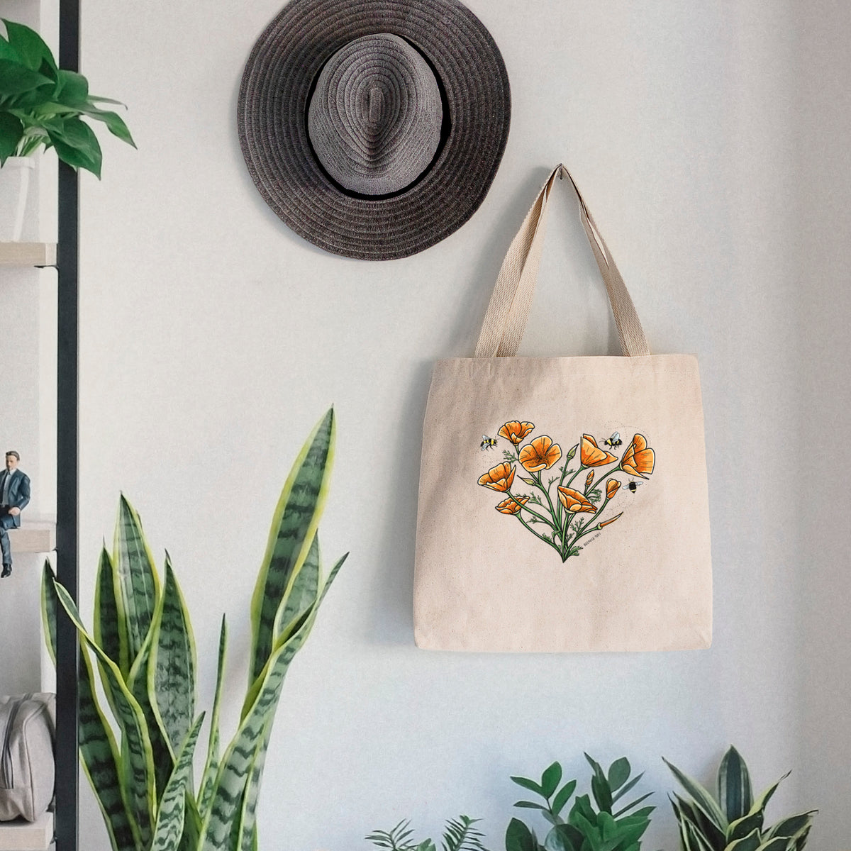 Canvas tote bag printed with a full-color bouquet of orange California poppies arranged in a heart shape, with small bees, hanging on a wall hook below a hat, next to potted plants.