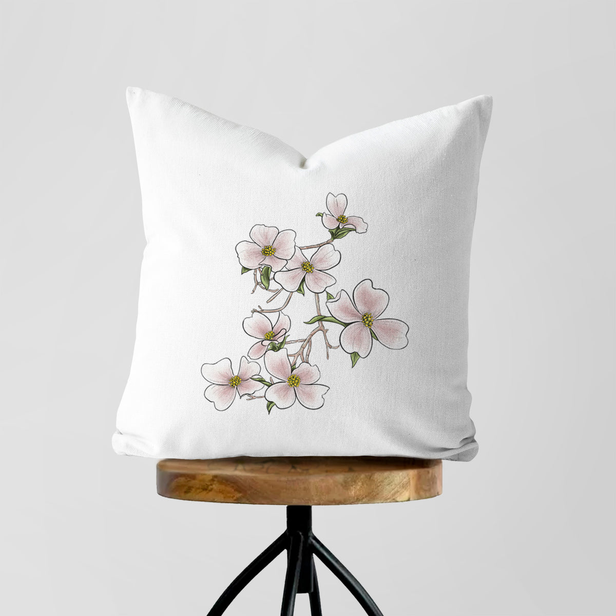 White pillow cover printed with a full-color illustration of pink and white dogwood blossoms on a branch, on a wooden stool.