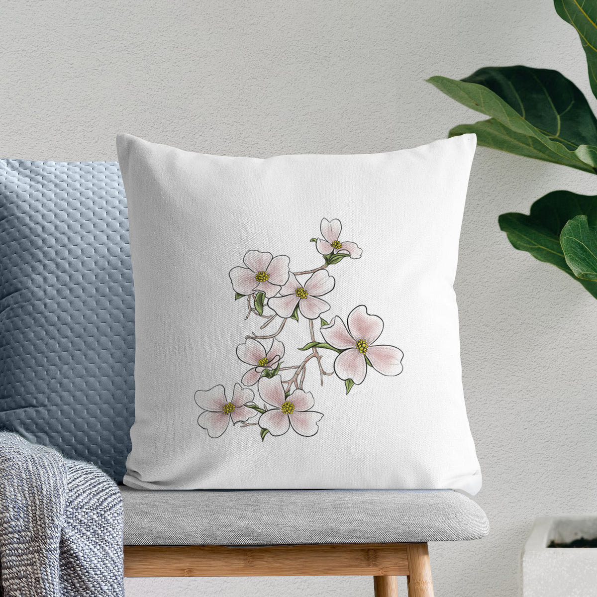 White pillow cover printed with a full-color illustration of pink and white dogwood blossoms on a branch, on a bench beside plants.