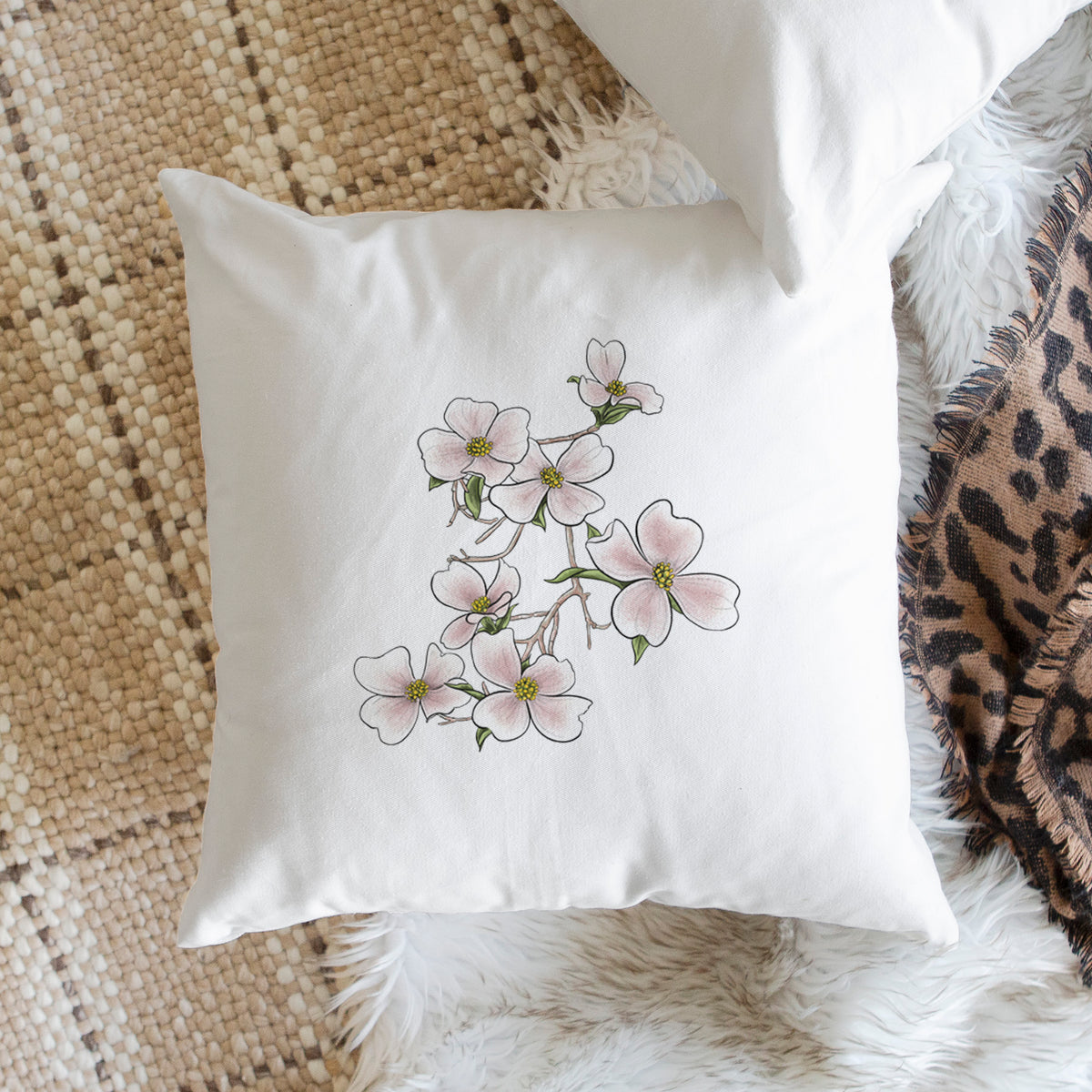 White pillow cover printed with a full-color illustration of pink and white dogwood blossoms on a branch, on a woven chair.