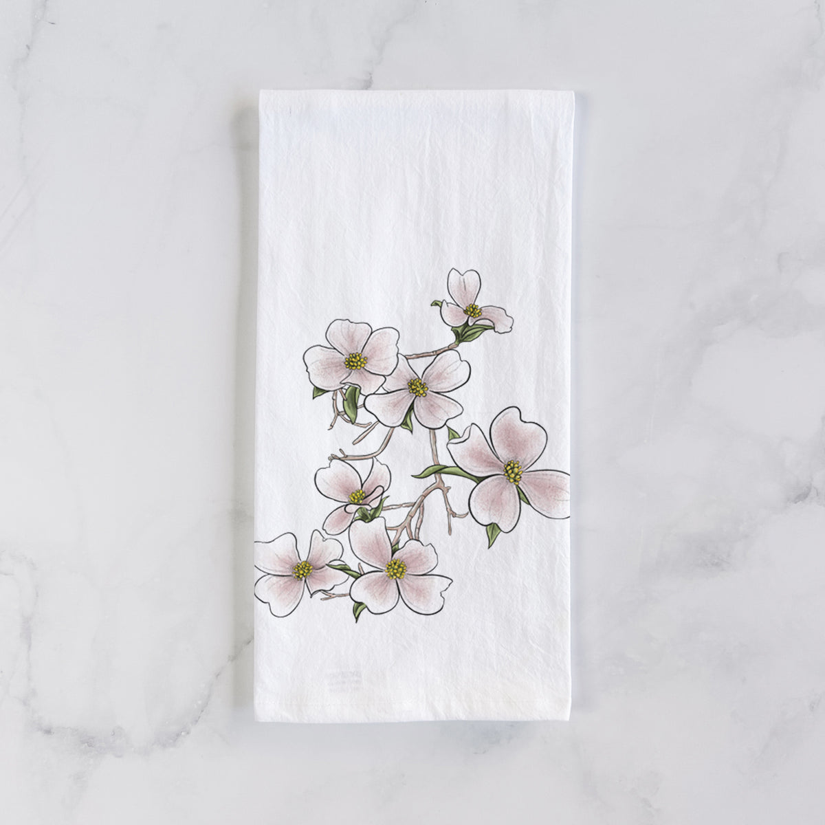 White cotton tea towel laid flat on marble, printed with a full-color illustration of pink and white dogwood blossoms on a branch.