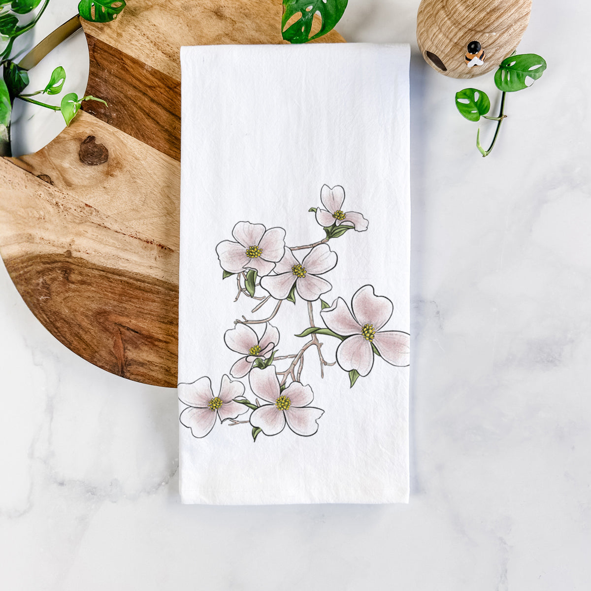 White cotton tea towel printed with a full-color illustration of pink and white dogwood blossoms on a branch, on a marble counter beside a round wooden cutting board and greenery.