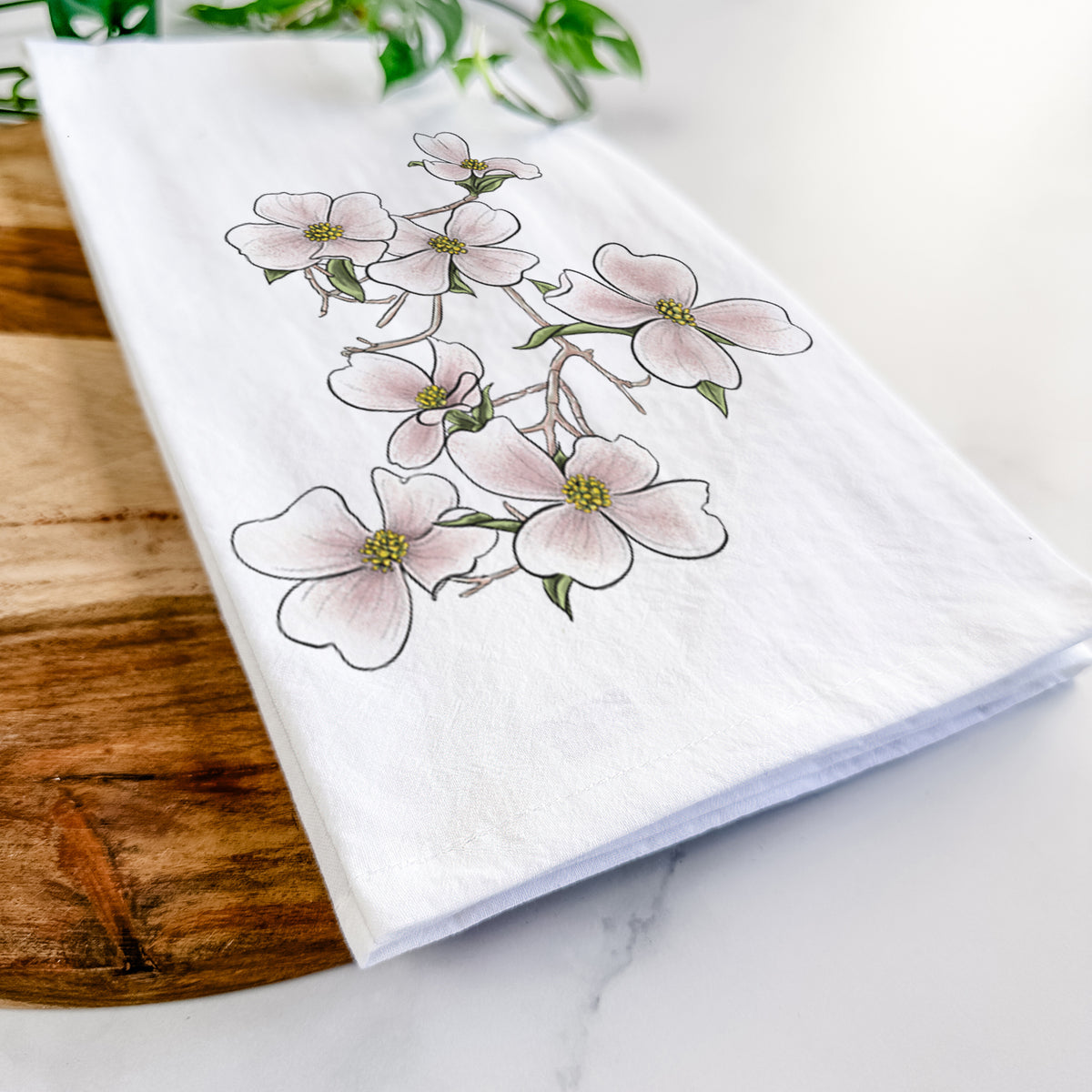 Close-up of a folded white tea towel on a wooden board, showing a full-color illustration of pink and white dogwood blossoms on a branch.
