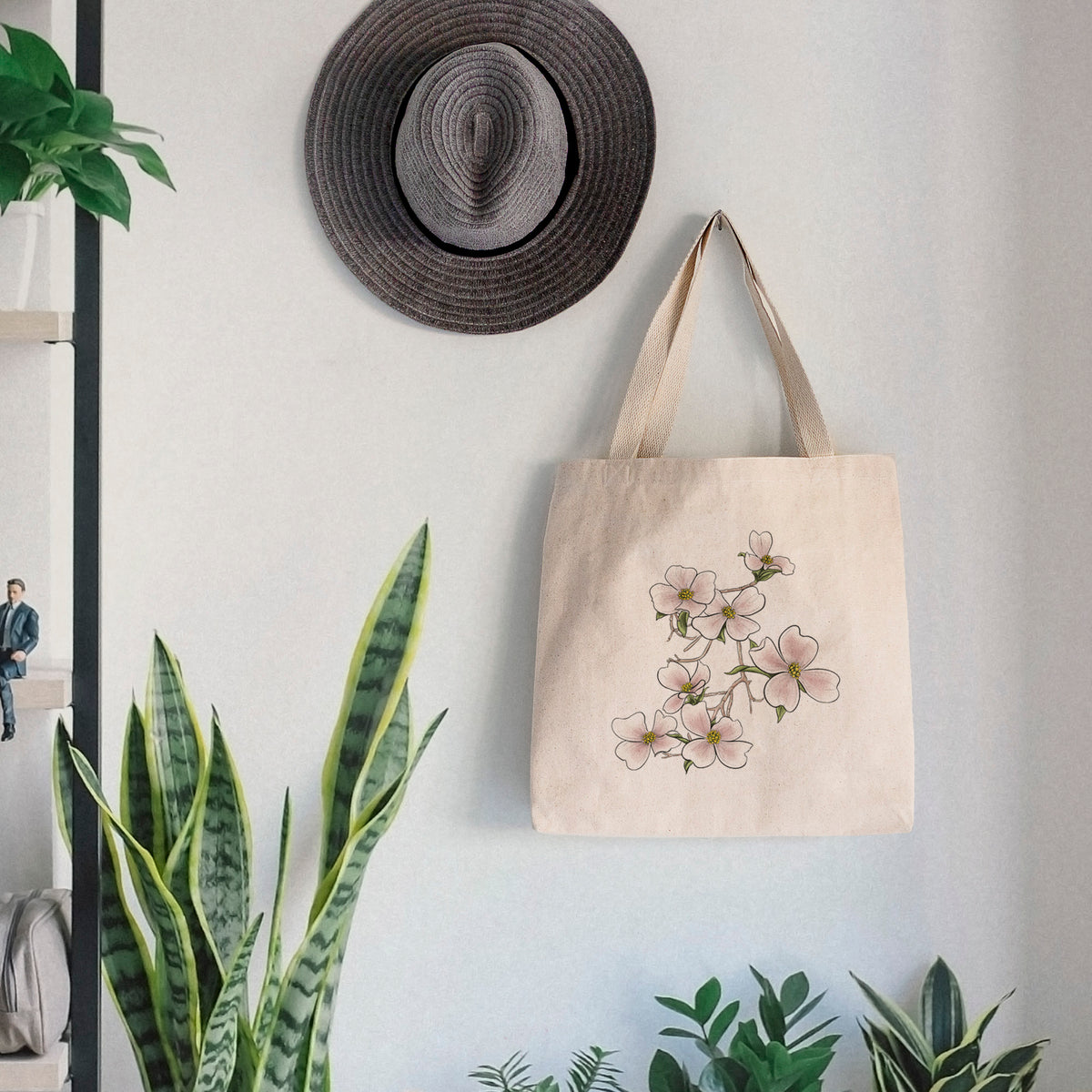 Canvas tote bag printed with a full-color illustration of pink and white dogwood blossoms on a branch, hanging on a wall hook below a hat, next to potted plants.