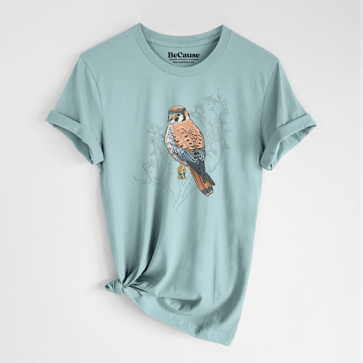 Dusty blue lightweight cotton t-shirt laid flat with the sleeves rolled, printed with a full-color illustration of an American kestrel perched among pale branches.