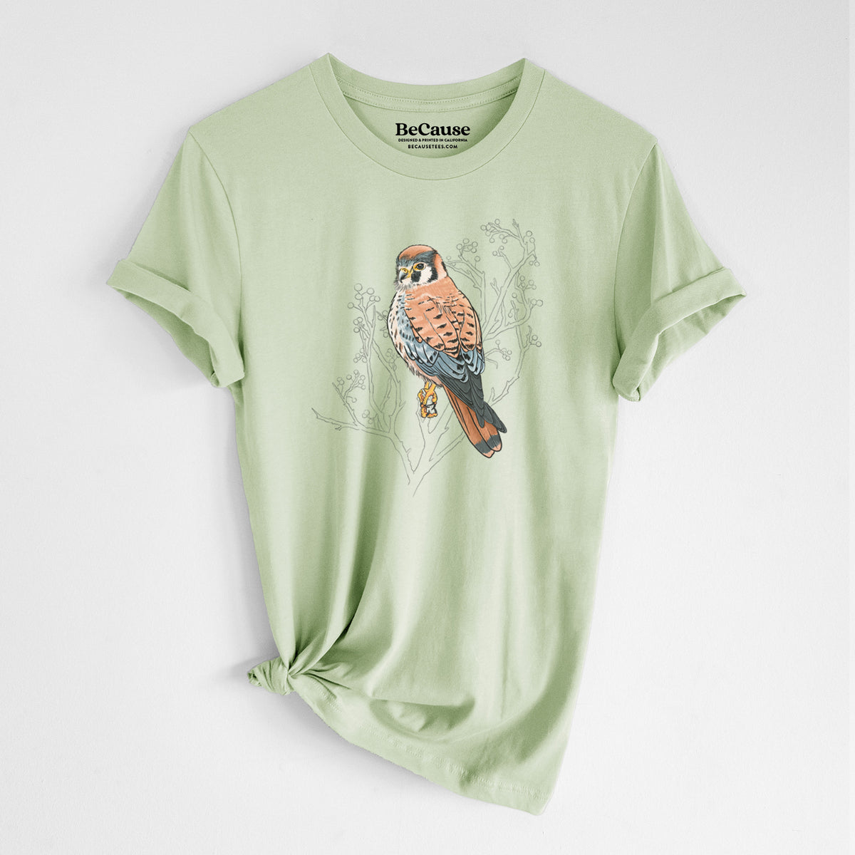 Spring green lightweight cotton t-shirt laid flat with the sleeves rolled, printed with a full-color illustration of an American kestrel perched among pale branches.