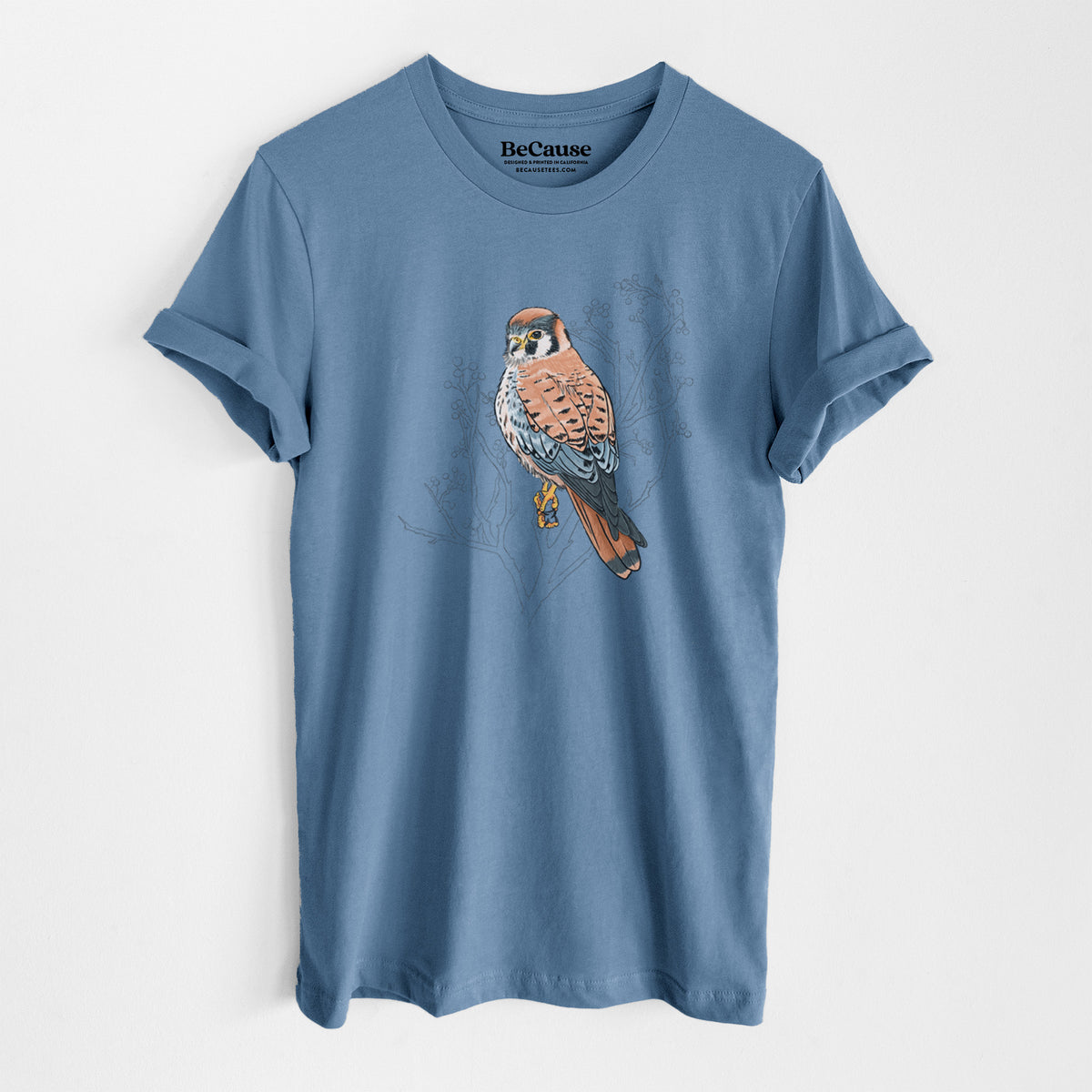 Steel blue lightweight cotton t-shirt laid flat, printed with a full-color illustration of an American kestrel perched among pale branches.