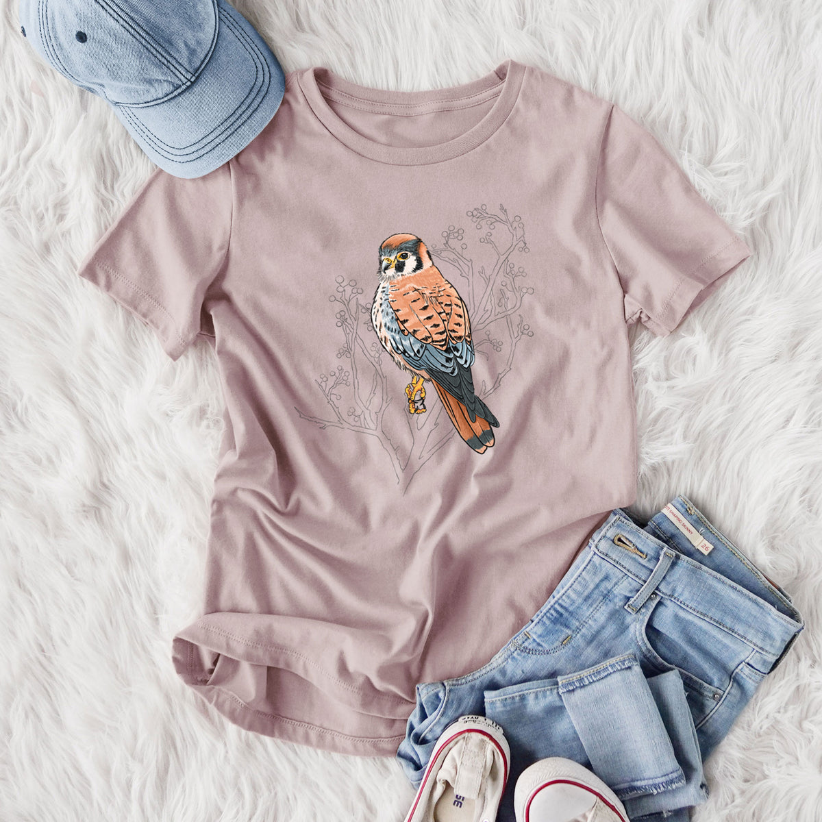 Women's relaxed fit cotton t-shirt printed with a full-color illustration of an American kestrel perched among pale branches, styled flat on a white faux fur rug with jeans.