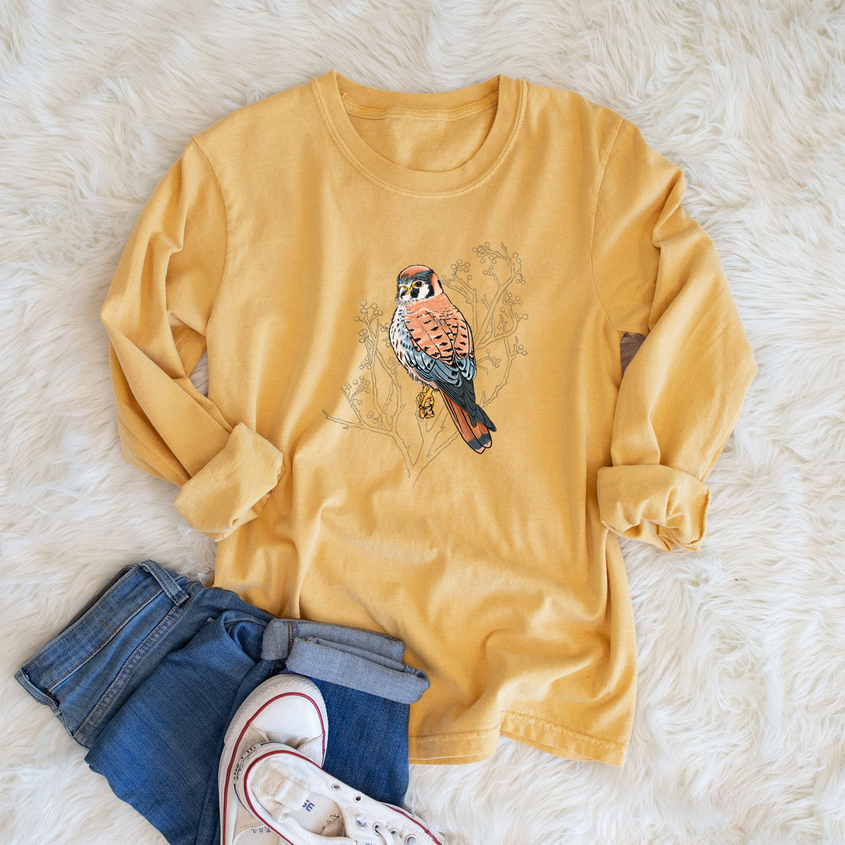 Golden wheat yellow Comfort Colors long sleeve shirt printed with a full-color illustration of an American kestrel perched among pale branches, shown in a lifestyle photo.