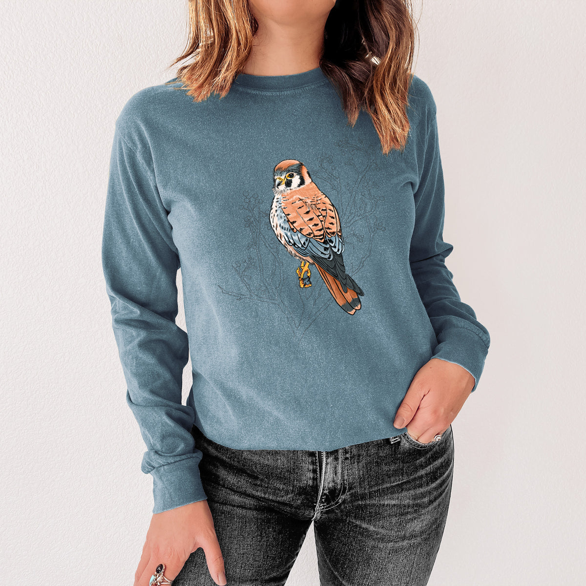 Steel blue Comfort Colors long sleeve shirt printed with a full-color illustration of an American kestrel perched among pale branches, shown in a lifestyle photo.