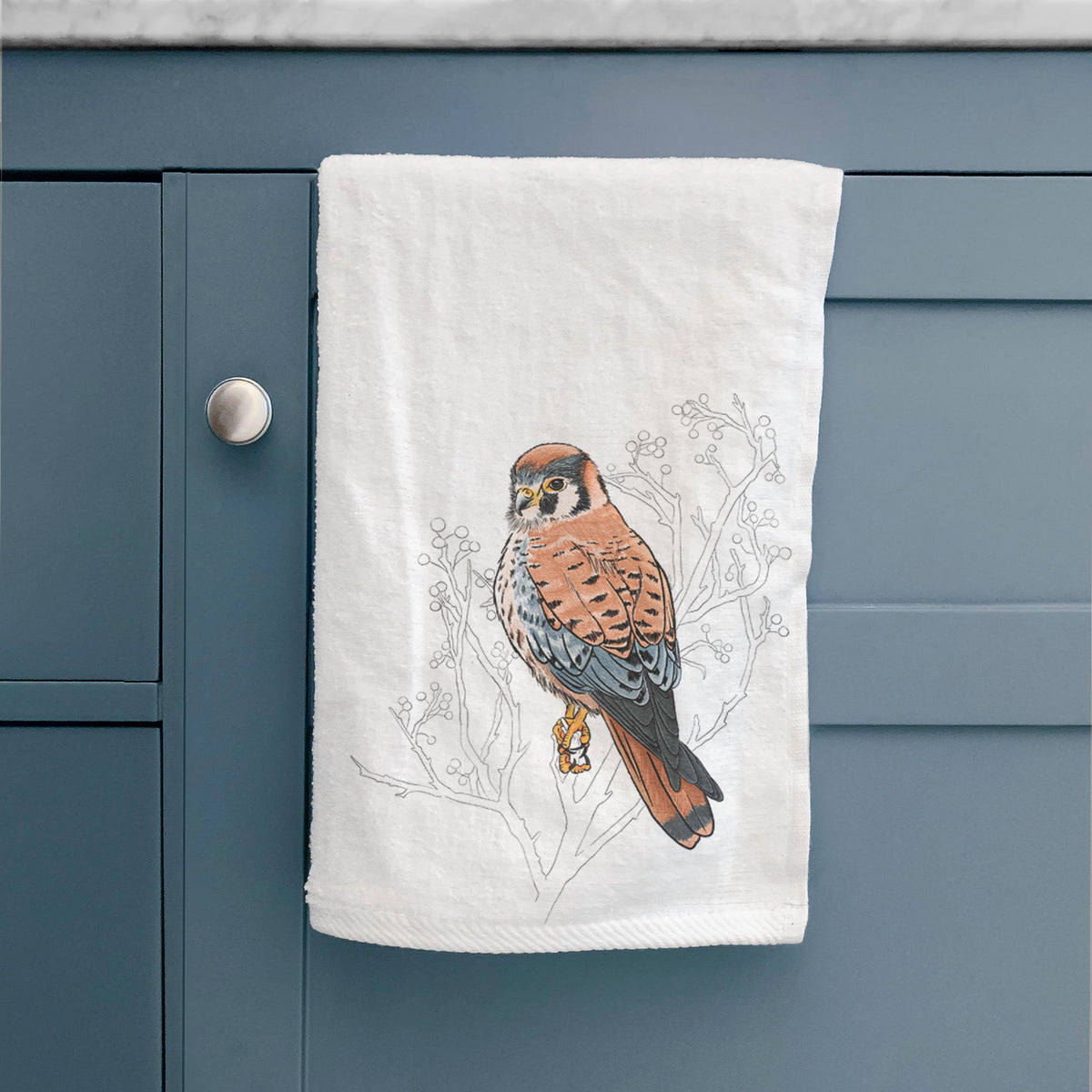White hand towel printed with a full-color illustration of an American kestrel perched among pale branches, hanging on a cabinet door.