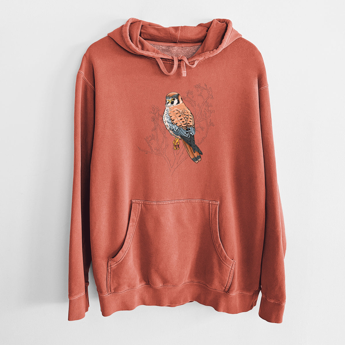 Amber orange pigment dyed hoodie laid flat, printed with a full-color illustration of an American kestrel perched among pale branches.