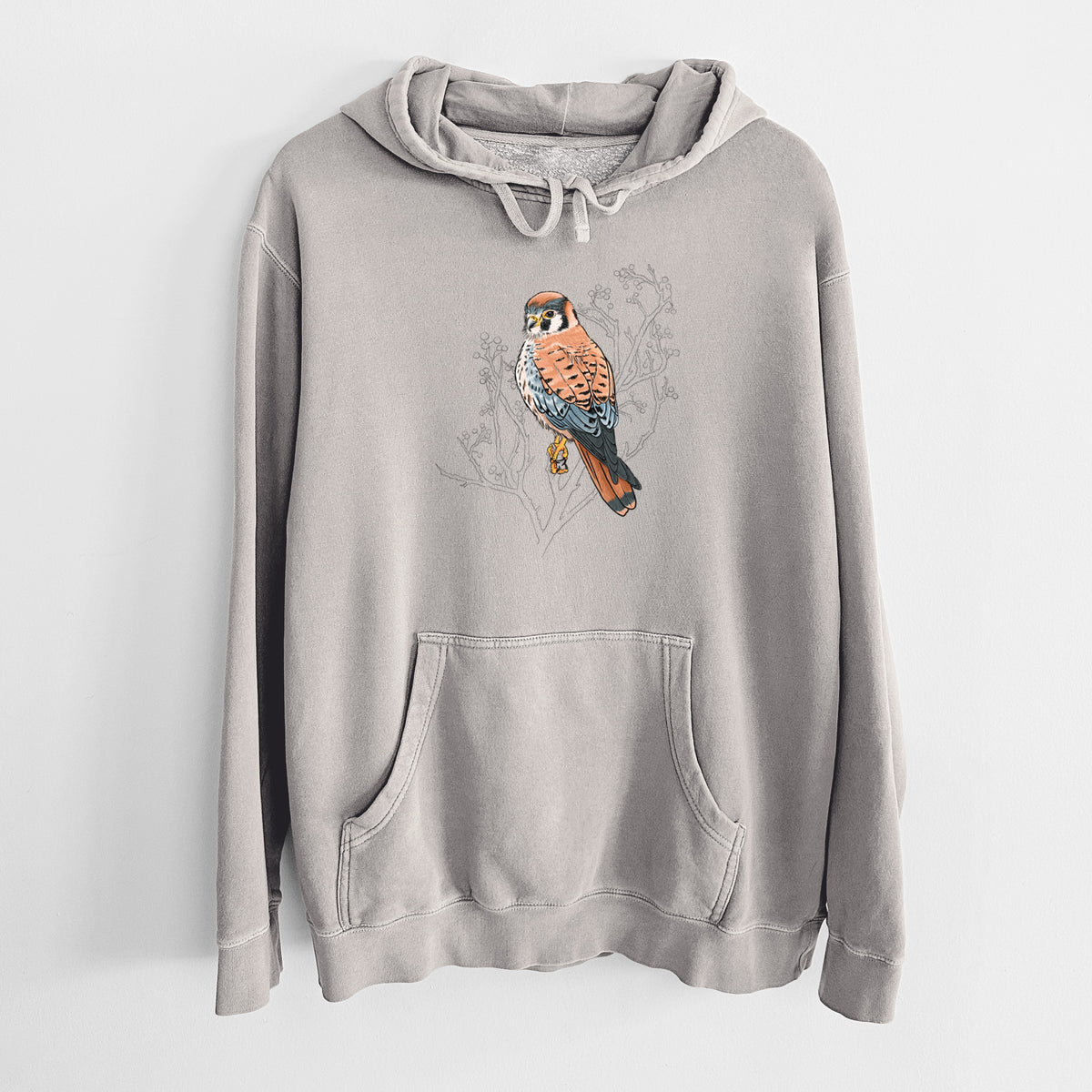 Cement grey pigment dyed hoodie laid flat, printed with a full-color illustration of an American kestrel perched among pale branches.