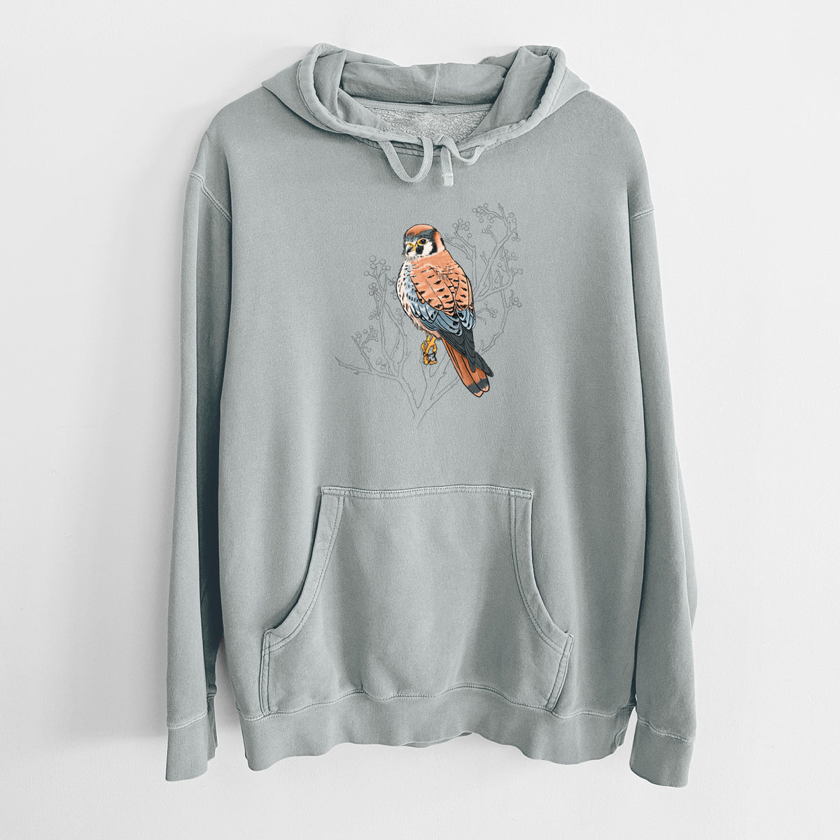 Sage green pigment dyed hoodie laid flat, printed with a full-color illustration of an American kestrel perched among pale branches.