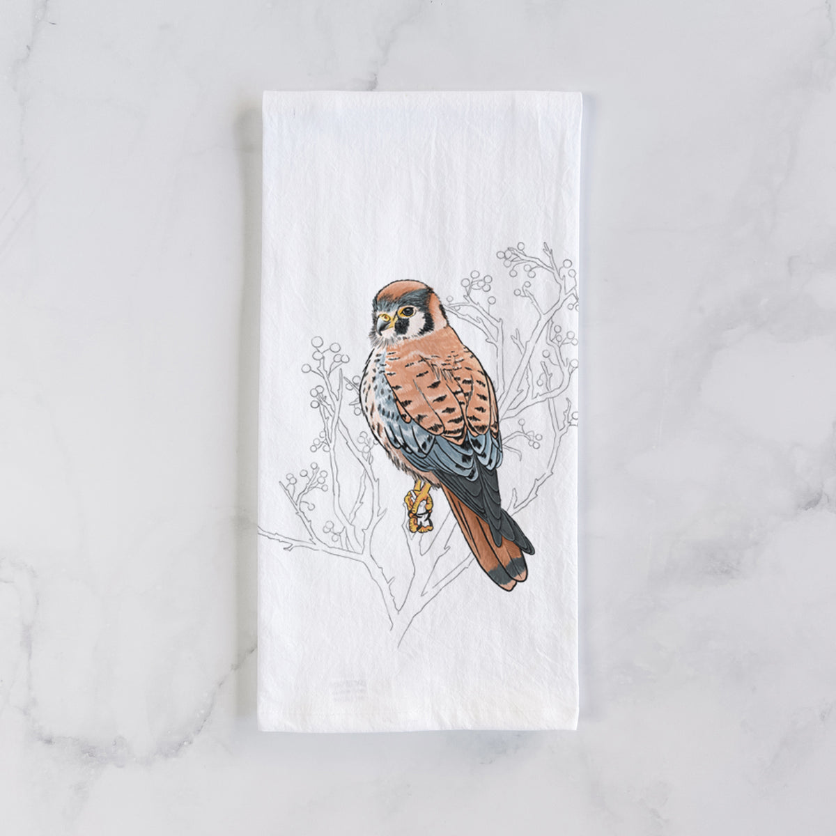 White cotton tea towel laid flat on marble, printed with a full-color illustration of an American kestrel perched among pale branches.