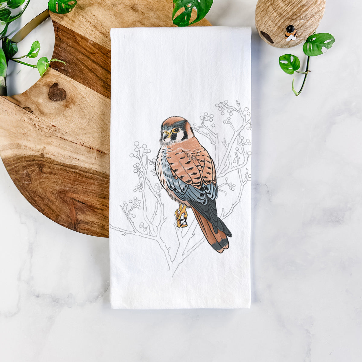 White cotton tea towel printed with a full-color illustration of an American kestrel perched among pale branches, on a marble counter beside a round wooden cutting board and greenery.
