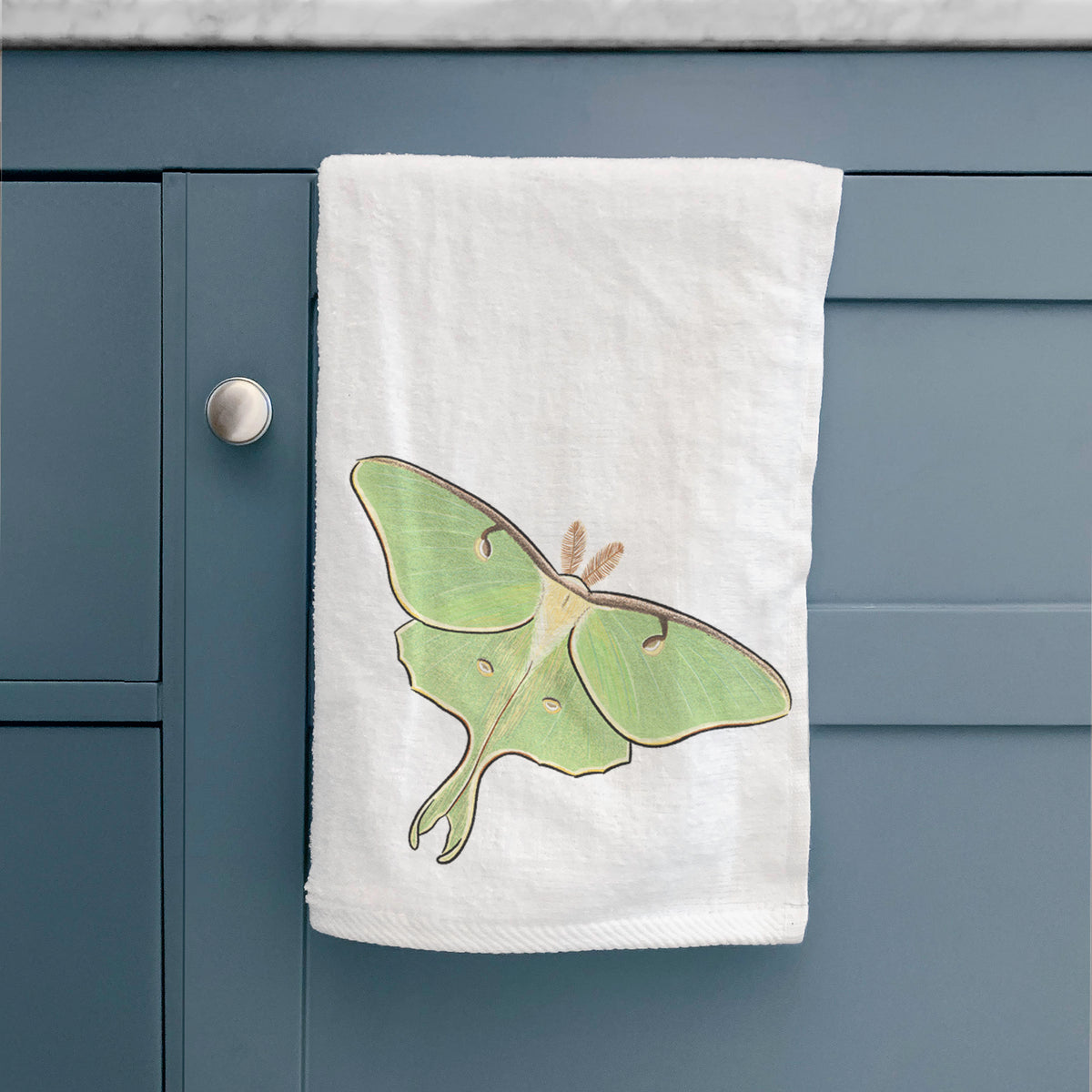 White hand towel printed with a full-color illustration of a pale green luna moth with long tails on its wings, hanging on a cabinet door.