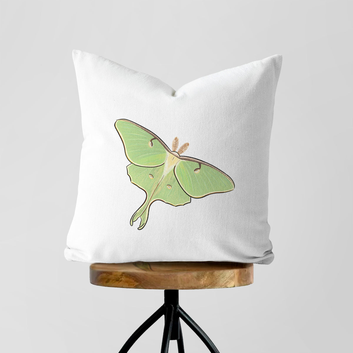 White pillow cover printed with a full-color illustration of a pale green luna moth with long tails on its wings, on a wooden stool.