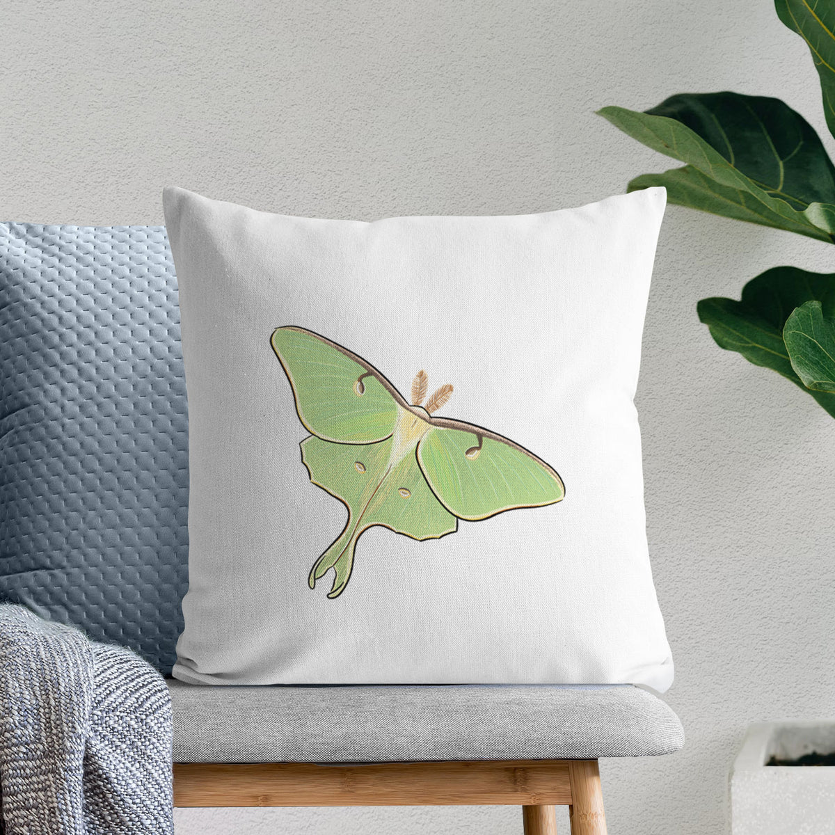 White pillow cover printed with a full-color illustration of a pale green luna moth with long tails on its wings, on a bench beside plants.