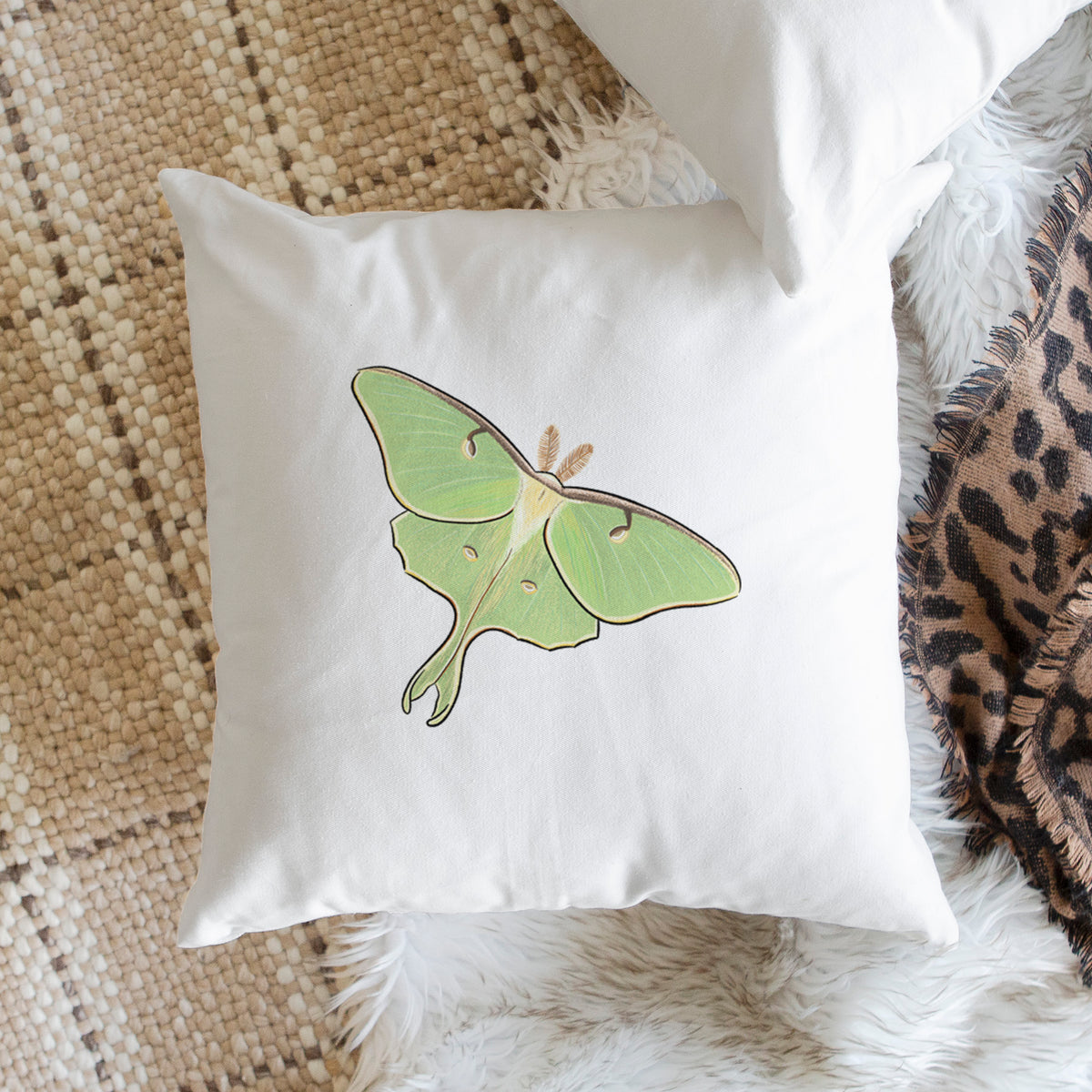 White pillow cover printed with a full-color illustration of a pale green luna moth with long tails on its wings, on a woven chair.
