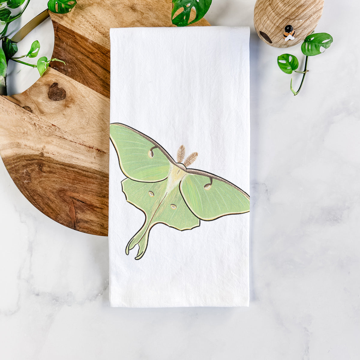 White cotton tea towel printed with a full-color illustration of a pale green luna moth with long tails on its wings, on a marble counter beside a round wooden cutting board and greenery.