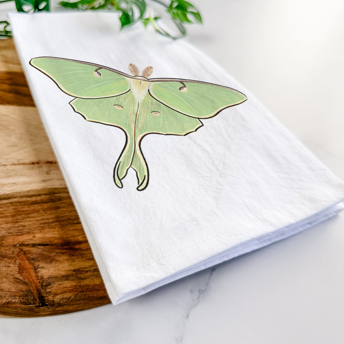 Close-up of a folded white tea towel on a wooden board, showing a full-color illustration of a pale green luna moth with long tails on its wings.