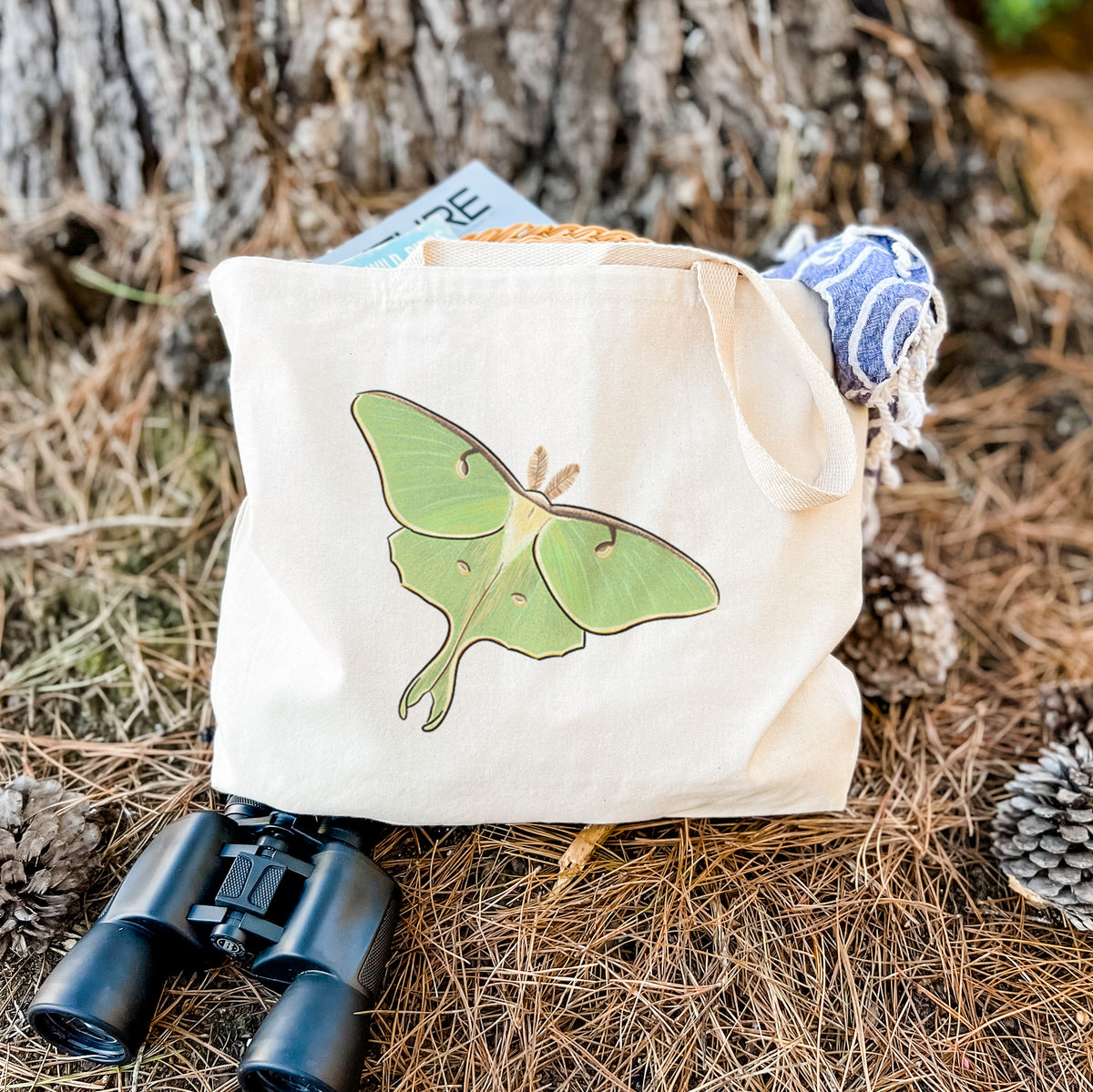 Canvas tote bag printed with a full-color illustration of a pale green luna moth with long tails on its wings, resting on dry grass beside a pair of binoculars.