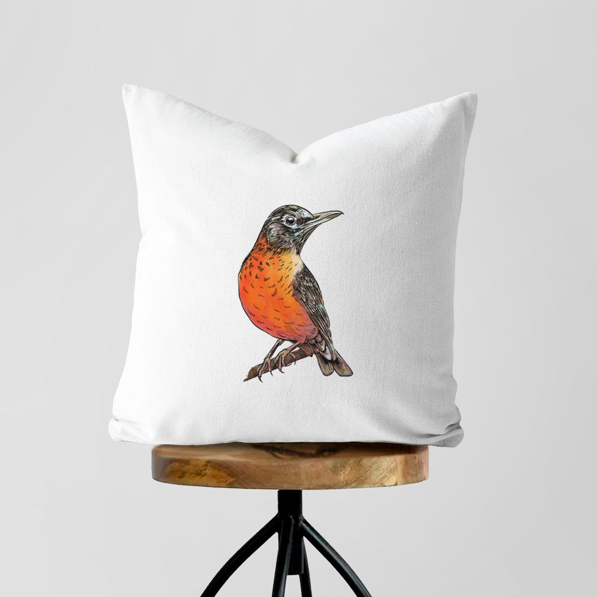 White pillow cover printed with a full-color illustration of an American robin with an orange breast perched on a branch, on a wooden stool.