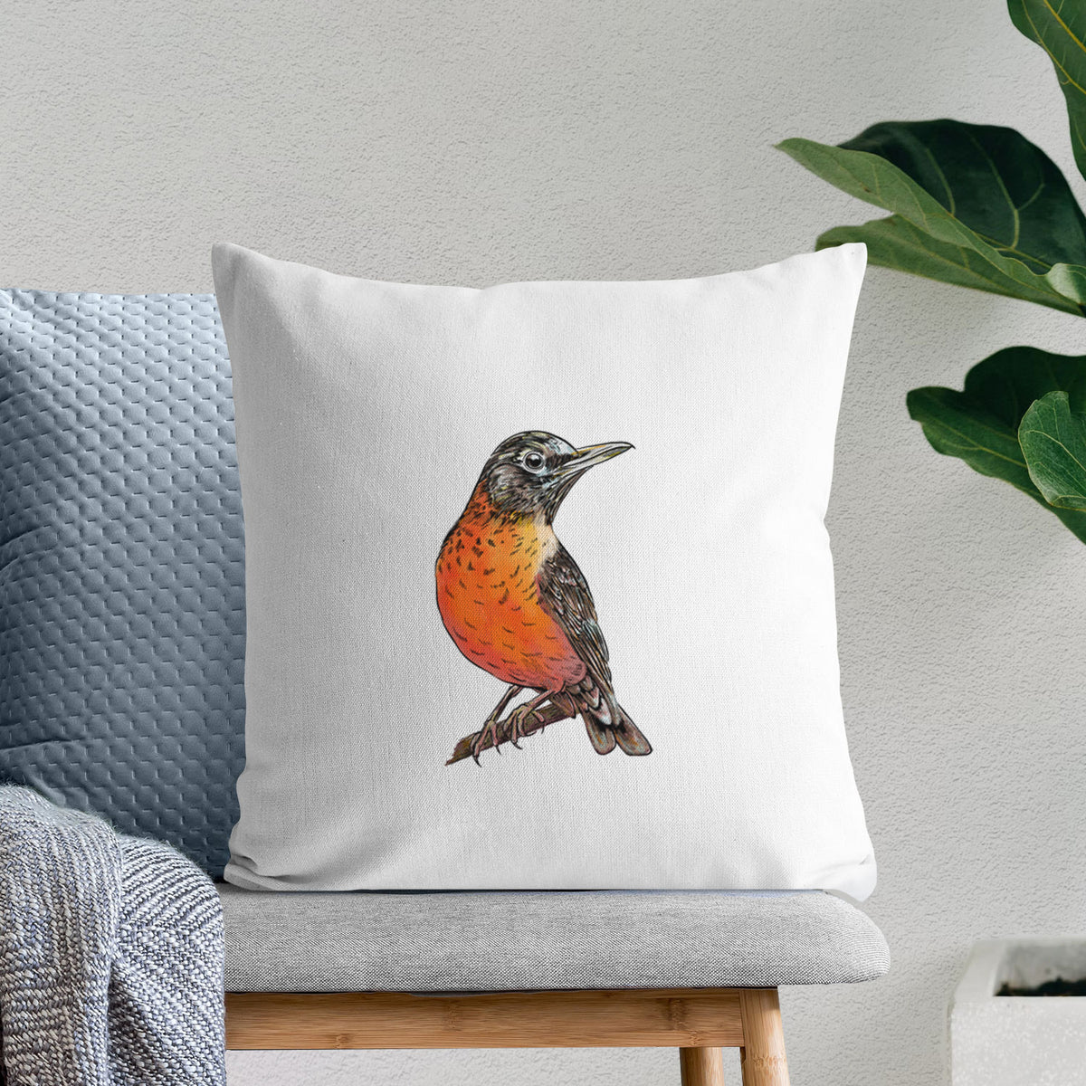 White pillow cover printed with a full-color illustration of an American robin with an orange breast perched on a branch, on a bench beside plants.