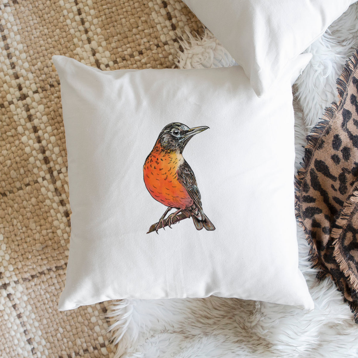 White pillow cover printed with a full-color illustration of an American robin with an orange breast perched on a branch, on a woven chair.