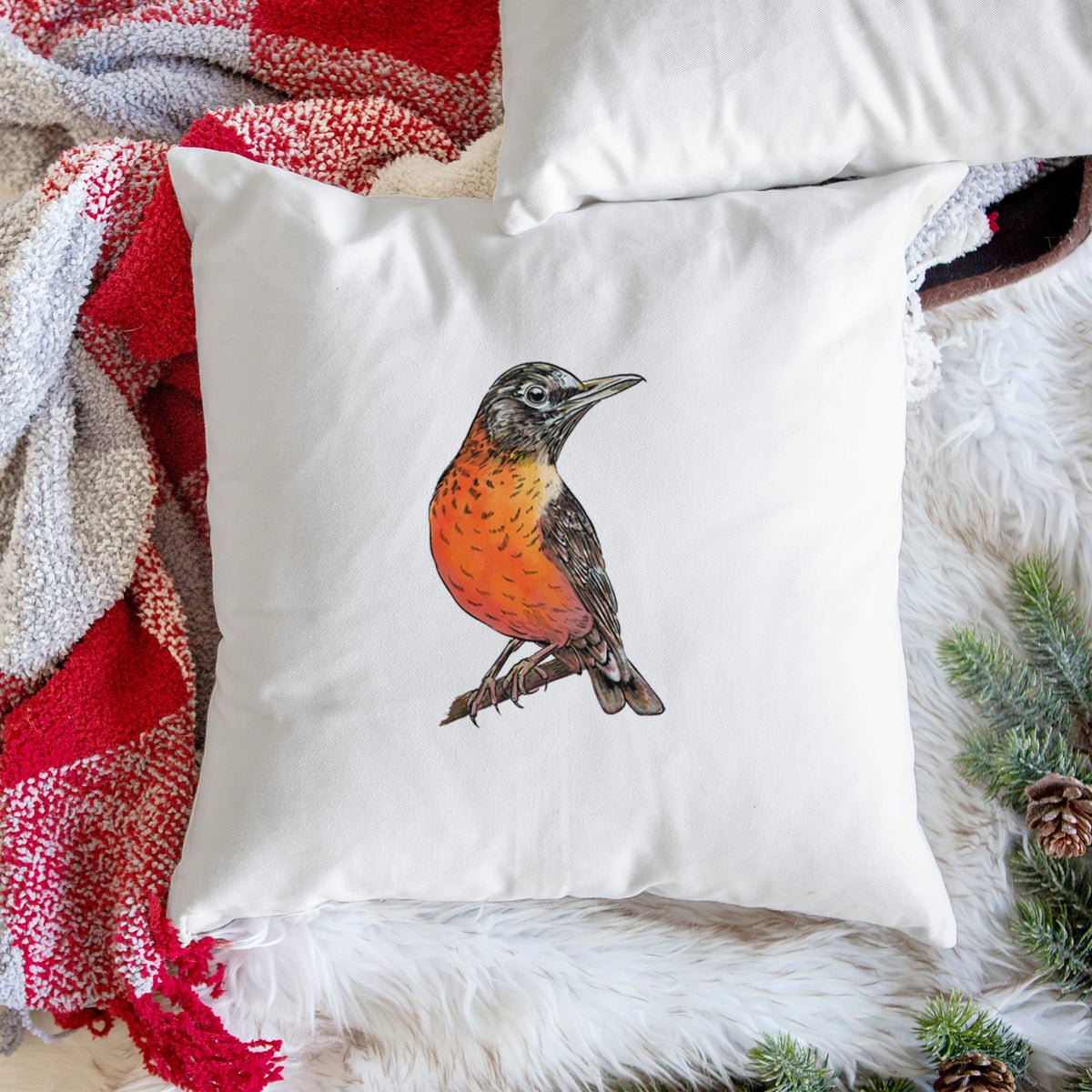 White pillow cover printed with a full-color illustration of an American robin with an orange breast perched on a branch, on a woven chair.