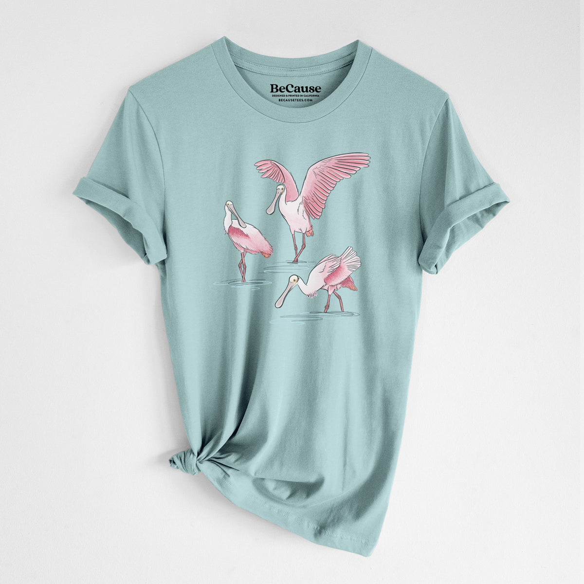 Dusty blue lightweight cotton t-shirt laid flat with the sleeves rolled, printed with a full-color illustration of three pink roseate spoonbills wading, one with its wings spread.