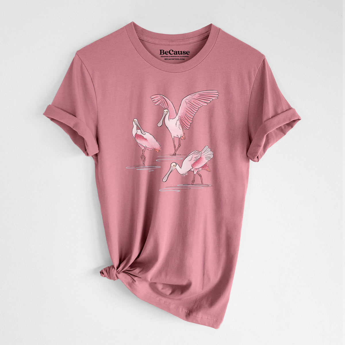 Mauve pink lightweight cotton t-shirt laid flat with the sleeves rolled, printed with a full-color illustration of three pink roseate spoonbills wading, one with its wings spread.