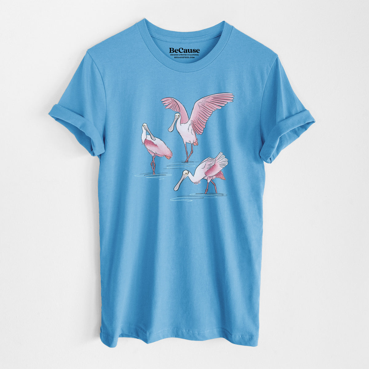 Ocean blue lightweight cotton t-shirt laid flat, printed with a full-color illustration of three pink roseate spoonbills wading, one with its wings spread.