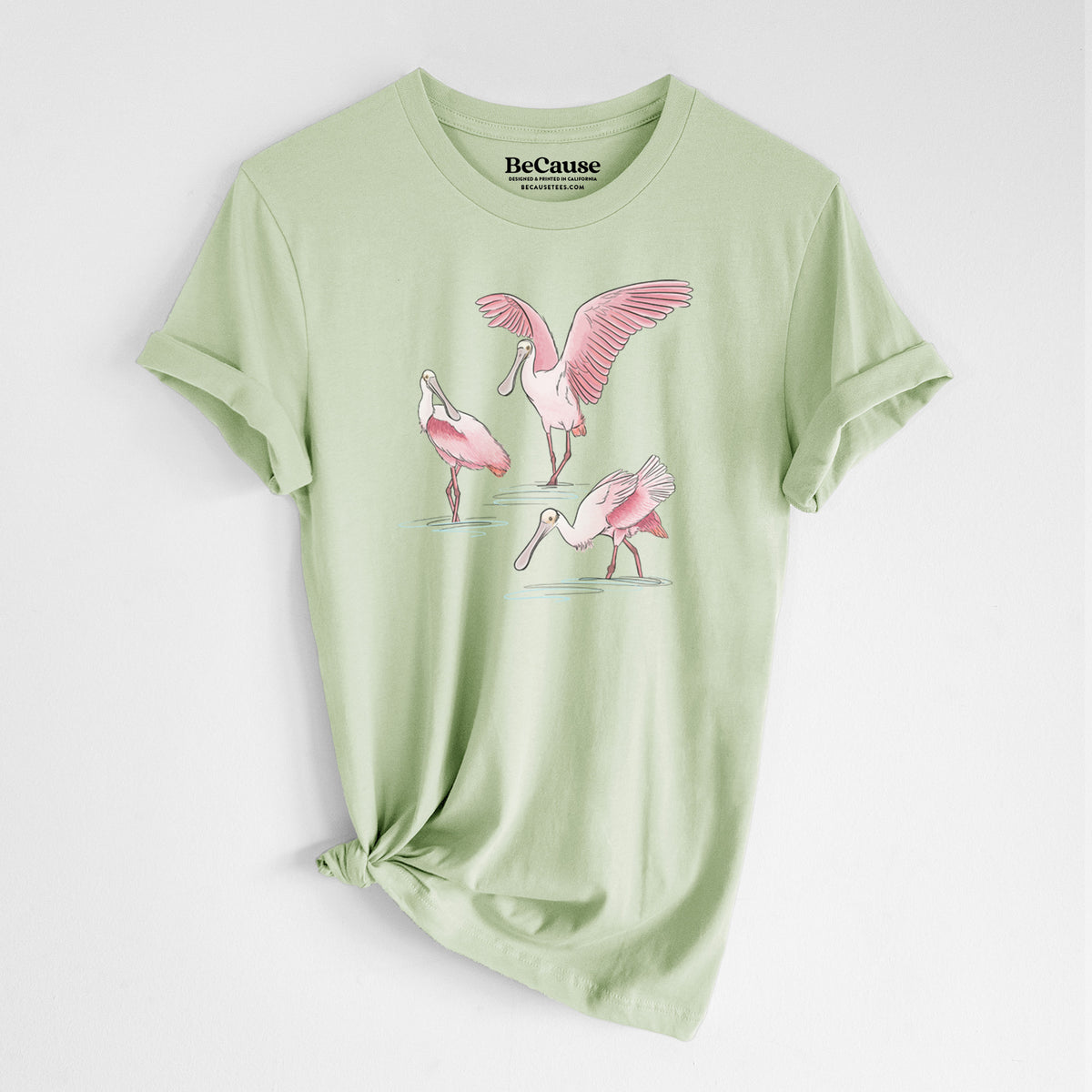 Spring green lightweight cotton t-shirt laid flat with the sleeves rolled, printed with a full-color illustration of three pink roseate spoonbills wading, one with its wings spread.