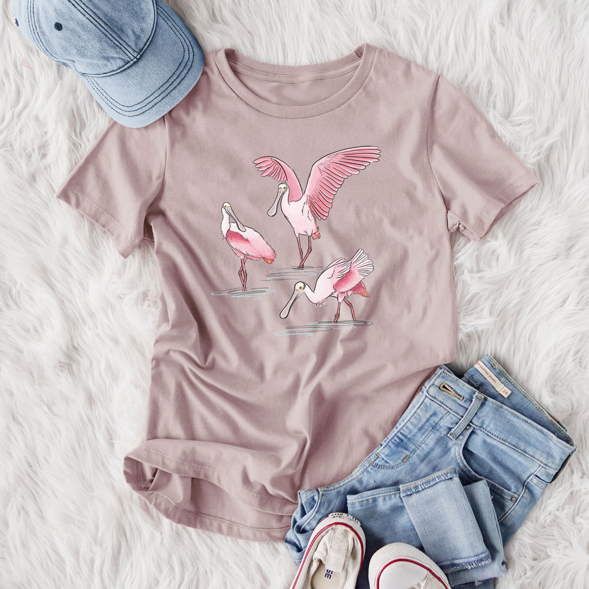 Women's relaxed fit cotton t-shirt printed with a full-color illustration of three pink roseate spoonbills wading, one with its wings spread, styled flat on a white faux fur rug with jeans.
