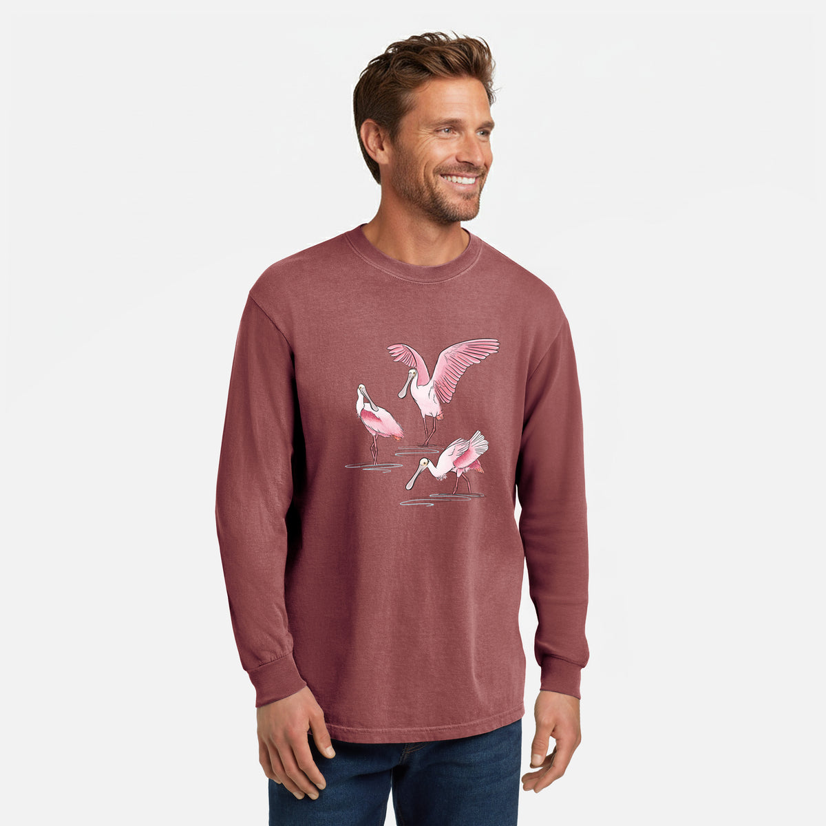 Brick red Comfort Colors long sleeve shirt printed with a full-color illustration of three pink roseate spoonbills wading, one with its wings spread, shown in a lifestyle photo.