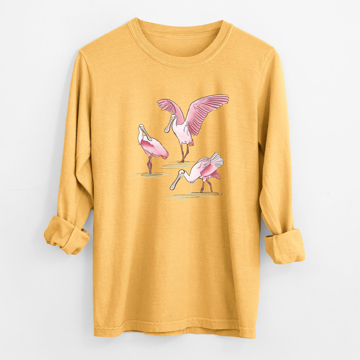 Golden wheat yellow Comfort Colors long sleeve shirt laid flat with the cuffs rolled, printed with a full-color illustration of three pink roseate spoonbills wading, one with its wings spread.