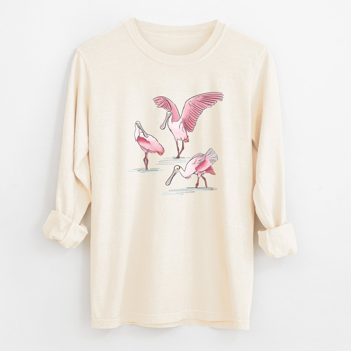 Ivory Comfort Colors long sleeve shirt laid flat with the cuffs rolled, printed with a full-color illustration of three pink roseate spoonbills wading, one with its wings spread.