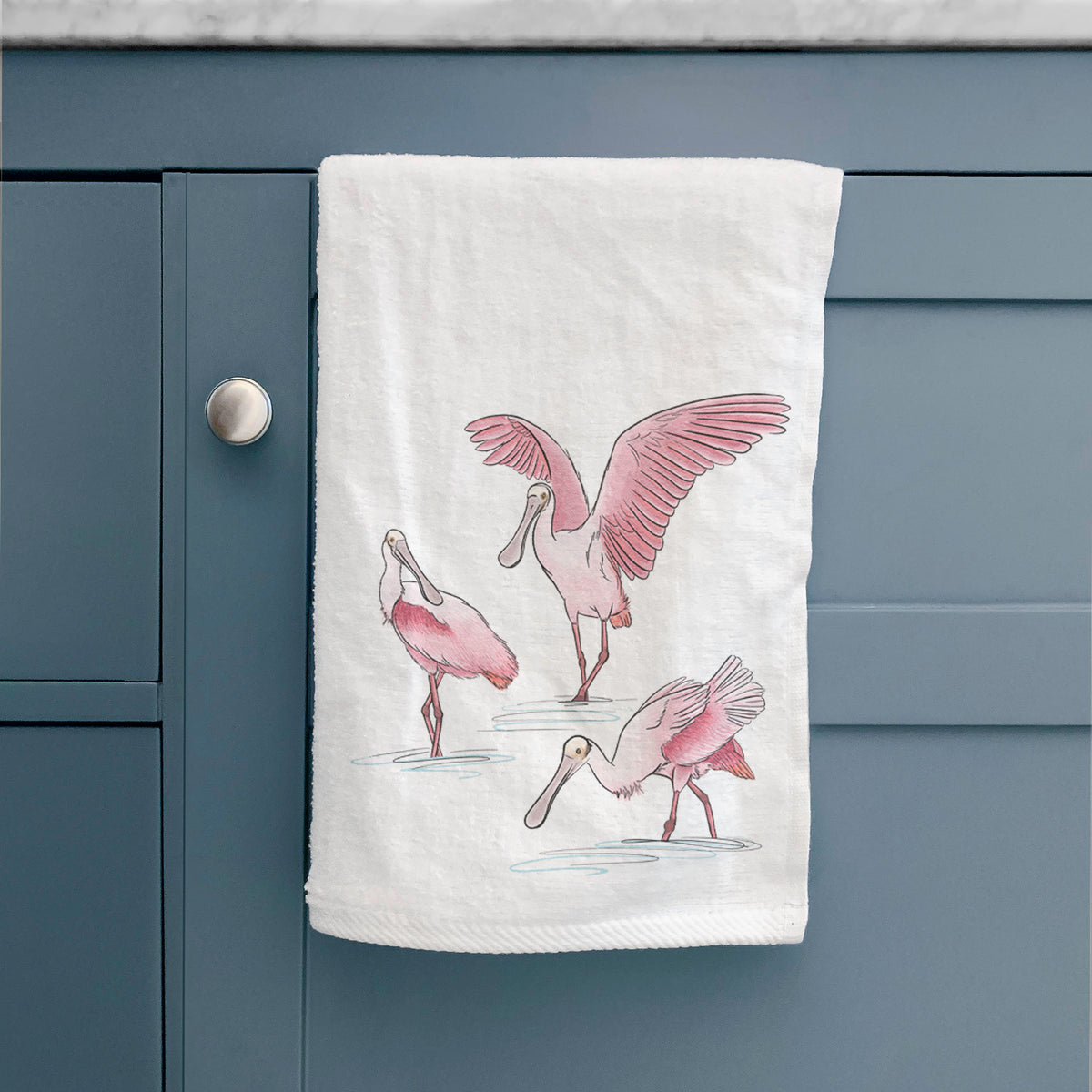 White hand towel printed with a full-color illustration of three pink roseate spoonbills wading, one with its wings spread, hanging on a cabinet door.